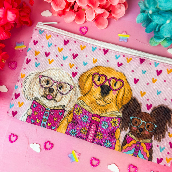 A rectangular zip pouch with a colorful illustration of three dogs wearing patterned sweaters, placed on a pink surface with pink flowers and small heart confetti around it.