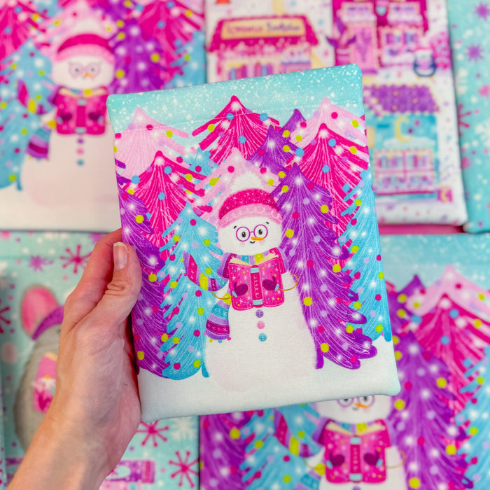 Hand holding a book sleeve featuring an illustration of a snowman holding a pink book in front of purple pink and blue trees.