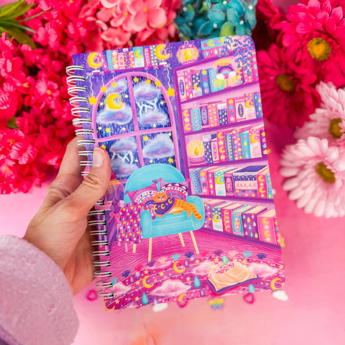 A hand holds a spiral-bound notebook with a colorful fantasy bookshelf illustration, surrounded by bright pink and teal flowers on a pink background.