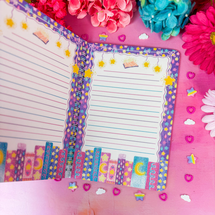 A colorful open planner or notepad with lined pages and decorative borders featuring stars, hearts, moons, and pastel patterns, styled on a pink surface with flowers and small paper accents around it.