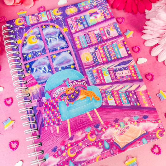 A spiral-bound notebook with a whimsical purple and blue library illustration, styled on a pink background with colorful flowers and decorative stickers.