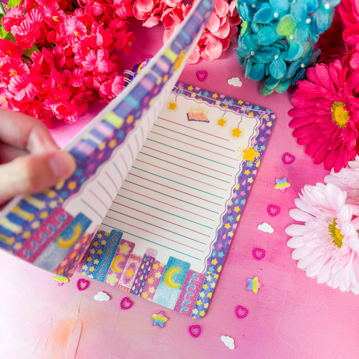 A hand holds an open lined notepad with a colorful patterned border, surrounded by bright pink and blue flowers and small decorative hearts on a pink surface.