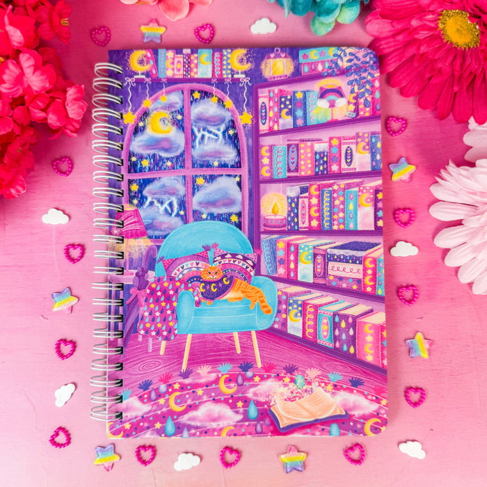 A spiral-bound notebook with a whimsical purple and blue library illustration, styled on a pink background with colorful flowers and decorative stickers.