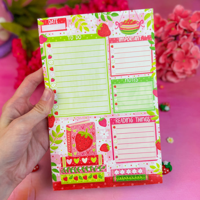 Notepad with strawberry design held by a hand against a pink background with flowers.