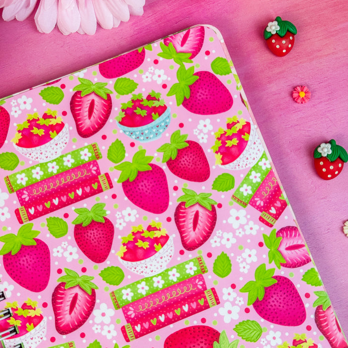 Pink notebook with strawberry pattern on a pink surface with flowers and decorative items.