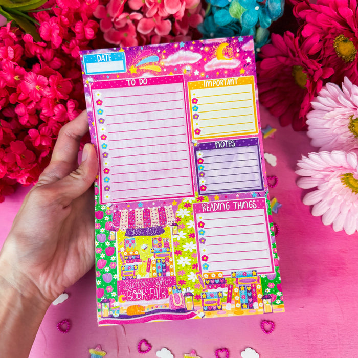 A hand holds a colorful planner notepad with sections for daily tasks, habit tracking, and reading themes, surrounded by bright pink and magenta flowers on a pink surface.