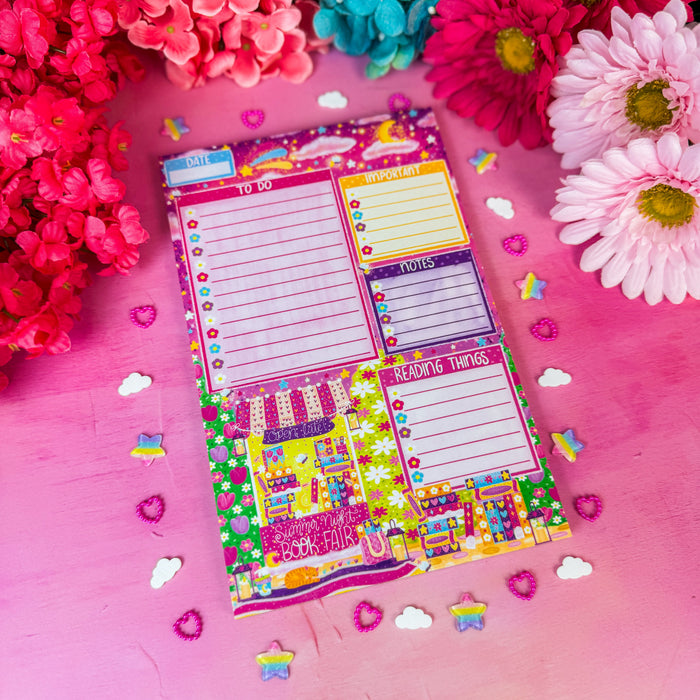 A colorful weekly planner notepad with lined boxes, notes sections, and cute illustrations, styled on a pink surface surrounded by bright flowers and small decorative stickers.