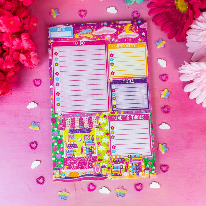A colorful planner notepad with lined sections, checkboxes, and decorative illustrations, photographed from above on a pink surface surrounded by bright flowers and small star and heart confetti.