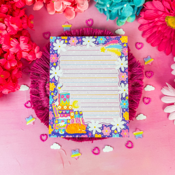 A colorful lined notepad with a floral border and cartoon character, styled on a pink surface surrounded by bright pink, red, and teal flowers.