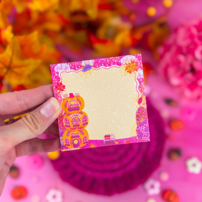 A hand holding sticky notes with 3 stacked pumpkins and pink, yellow, and red leaves and glitter around the edges. Against a bright pink background.
